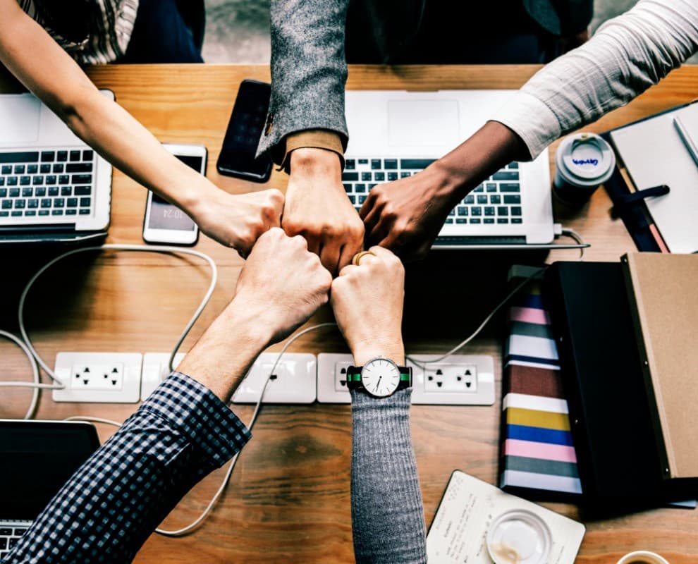Collaborators joining hands over a table with laptops, notebooks, and phones in a meeting